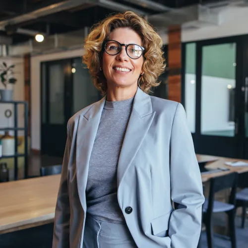 Smiling businesswoman with glasses wearing a gray suit standing confidently in a modern office.