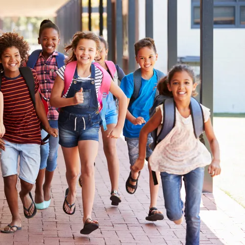 Group of diverse smiling children with backpacks running joyfully down a school hallway with lockers.