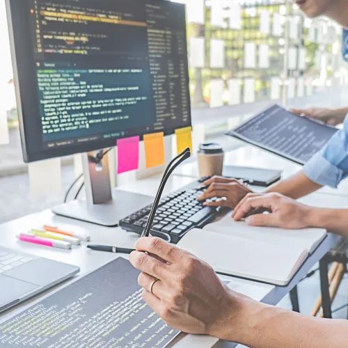 Two developers collaborating on coding projects with multiple screens and notes in a bright workspace.