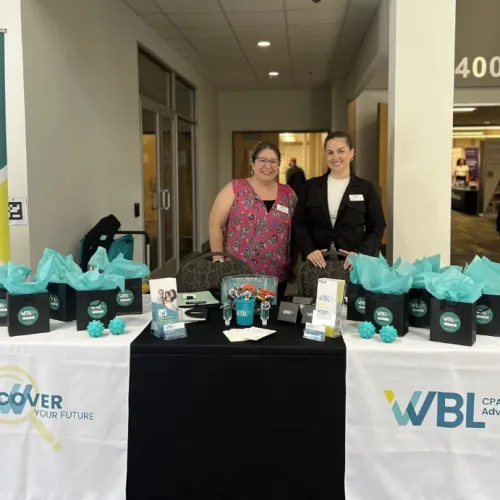Two women at a WBL CPAs and Advisors booth with promotional bags and materials at an event.