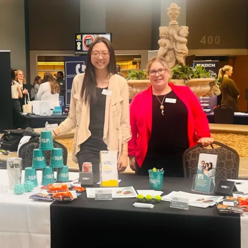 Two women stand behind a promotional table with branded cups and items at an indoor event with a fountain background.