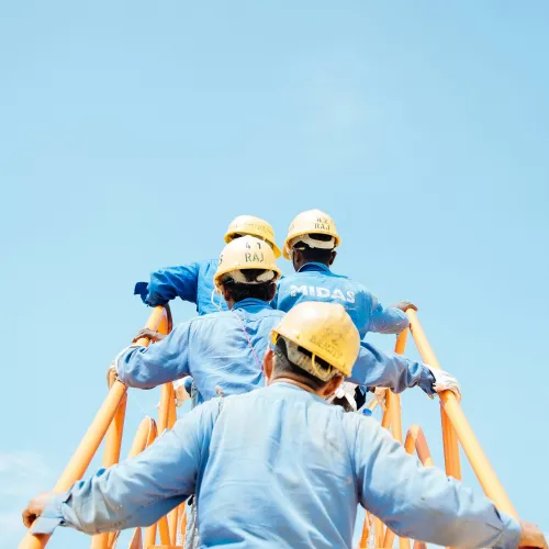 Construction workers in blue uniforms and yellow helmets climbing orange scaffolding against clear blue sky.