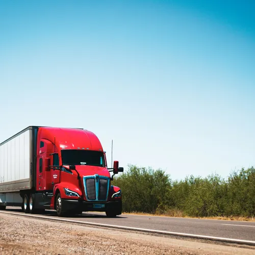 Red semi-truck driving on a highway with clear blue sky and green roadside vegetation on a sunny day
