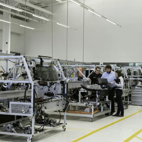 Engineers collaborating and reviewing plans on a laptop in a modern automotive manufacturing facility.
