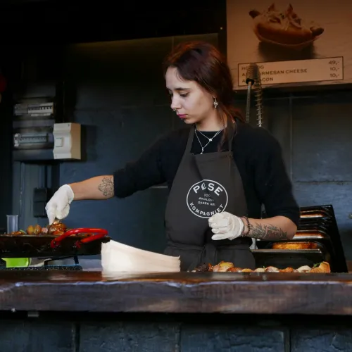Woman in apron cooking sausages at a food stall with menu boards and hotdog prices visible.