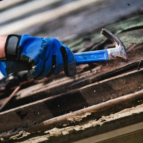 Worker wearing blue gloves uses a hammer to strip old wood and paint from a weathered surface outdoors.