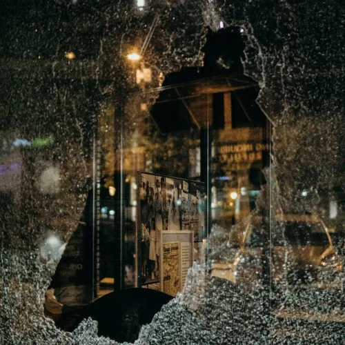 Shattered glass window frame with city lights and building reflections visible at night through the broken area.