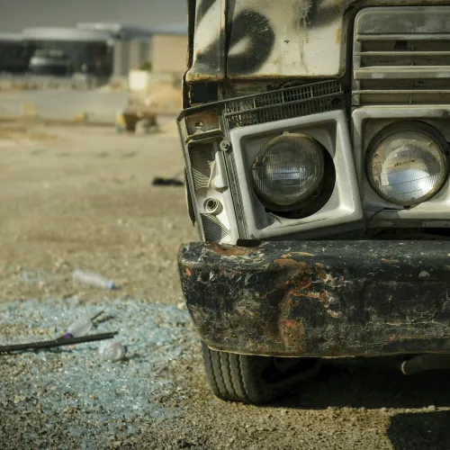 Close-up of a damaged vehicle front with broken headlights and rusty bumper on a gravel surface.