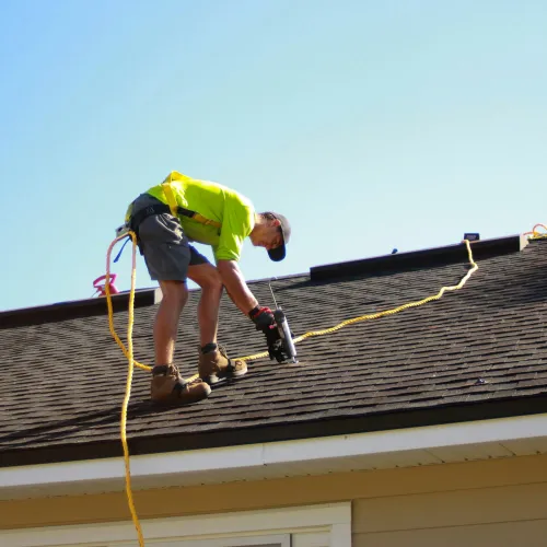 Roofer in bright shirt using nail gun to repair asphalt shingle roof on a sunny day.