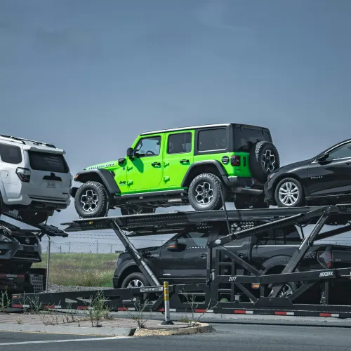 Green Jeep Wrangler and other vehicles loaded on a car carrier trailer under clear sky at a transport yard