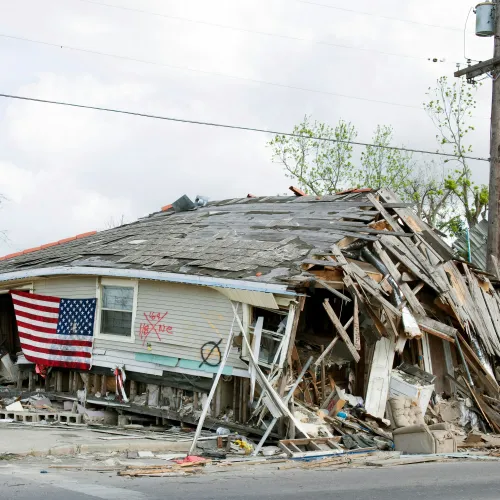 Severely damaged house with collapsed roof and walls, American flag hanging on the side amid debris and destruction