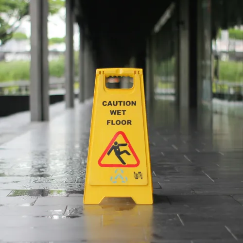 Yellow caution wet floor sign on a wet, dark tiled floor near glass windows in an outdoor corridor.