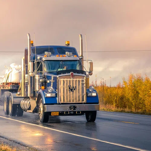 Blue semi-truck hauling covered cargo on wet road with autumn trees and cloudy sky in the background