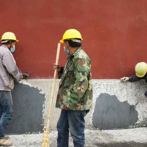 Three construction workers with helmets and masks repairing and chipping paint off a wall outdoors.