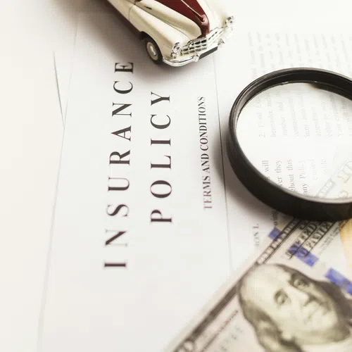 Close-up of an insurance policy document with a magnifying glass, toy car model, and US hundred-dollar bill