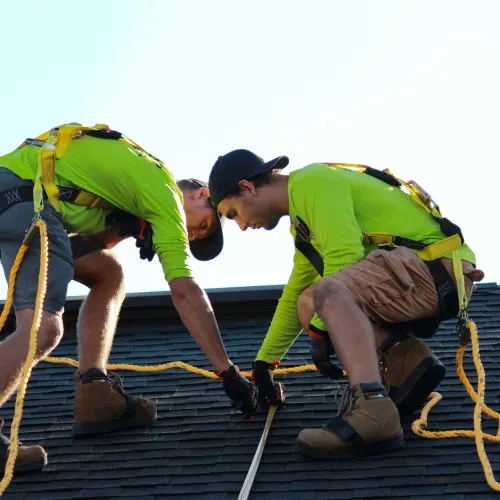 Two construction workers wearing safety harnesses measure a roof while kneeling on shingles under clear sky.