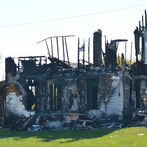 Burned remains of a house with charred walls and roof against a clear sky and green grass.
