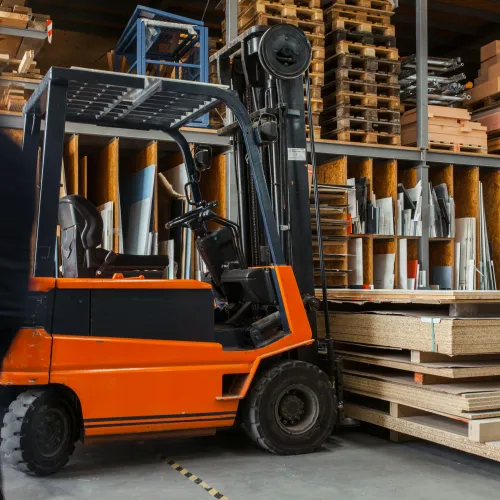 Orange forklift parked in a warehouse next to stacked plywood sheets and pallets with blurred worker nearby