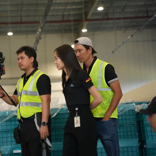 Three workers in high-visibility vests filming with a camera on a tripod inside an industrial warehouse.