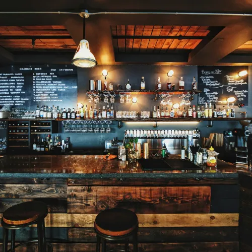 Cozy bar interior with wooden stools, hanging lights, bottles on shelves, and draft beer and coffee menus on chalkboards.