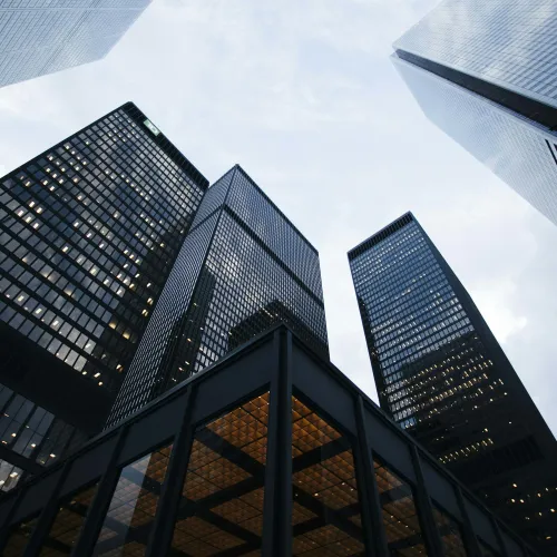 View looking up at tall modern glass skyscrapers with illuminated windows against a cloudy sky.