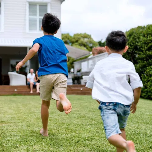 Two boys running barefoot on grass in a backyard with a modern house and adults seated in the background.