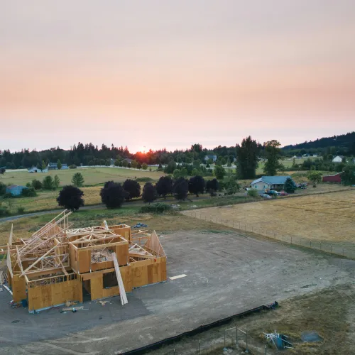 A house under construction at sunset in a rural area with fields and trees surrounding the site.