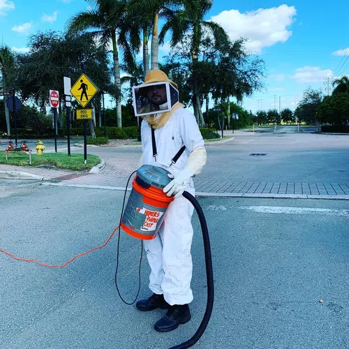 Person in white protective beekeeper suit holding a homemade vacuum labeled Homer's Pooter Bucket outdoors.
