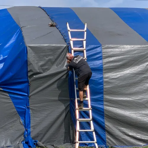 Man climbing aluminum ladder to adjust large blue and gray striped tarp or tent cover outdoors.