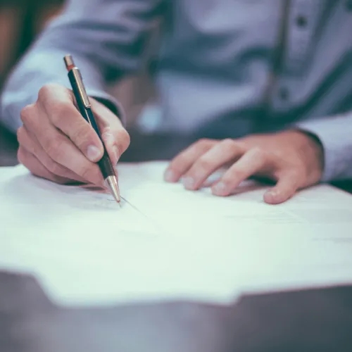 Person in blue shirt writing on white documents with a black pen at a dark desk.