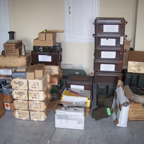 Stacks of labeled wooden and metal military ammunition boxes and crates stored indoors on a concrete floor.