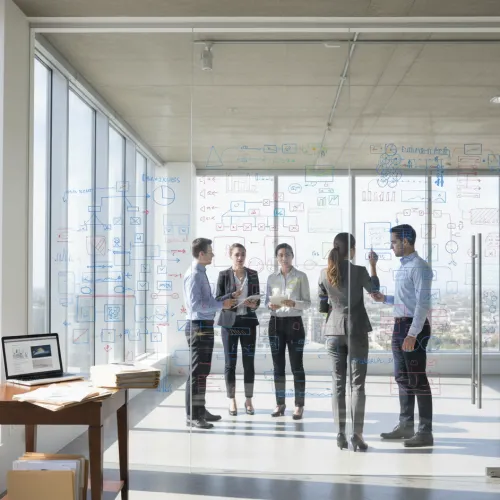 Five professionals brainstorming business strategy in a glass-walled office with flowcharts and diagrams drawn on the glass.