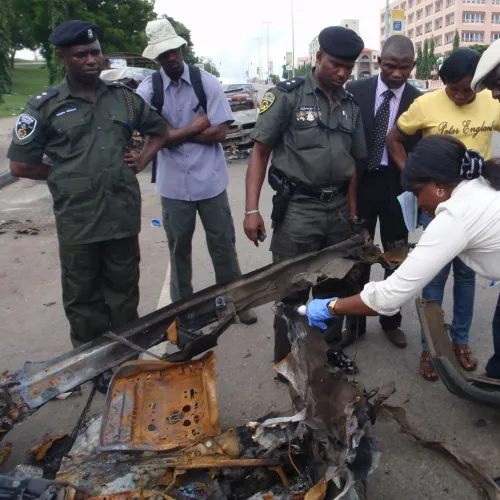 Police officers and civilians examining wreckage of a destroyed vehicle on a city road during investigation.