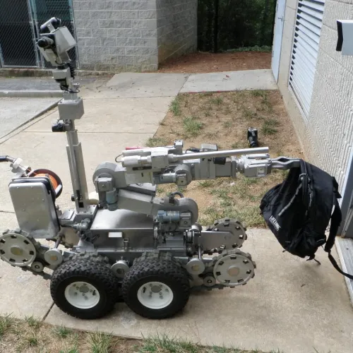 Robotic bomb disposal unit gripping a black backpack outside a building on concrete pavement.