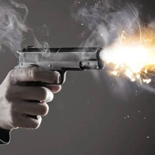 Hand holding a handgun firing with visible muzzle flash and smoke against a gray background