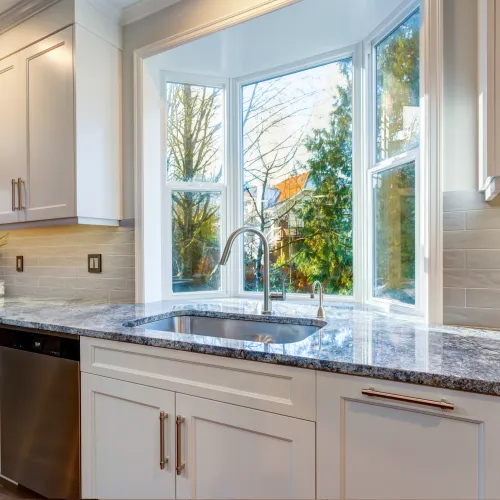 Bright modern kitchen with granite countertops, white cabinetry, stainless steel sink, and large bay window