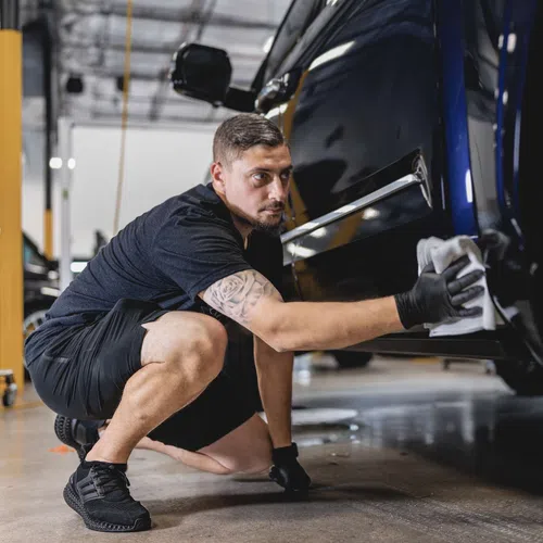 Man wearing black gloves carefully polishing a dark blue car in a well-lit garage.