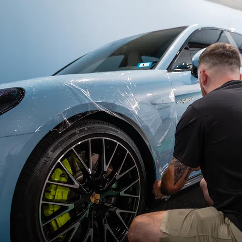 Man applying protective film on the side of a light blue Porsche sports car in a garage.