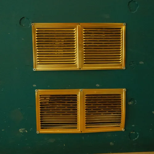 Two gold-colored ventilation grilles mounted on a green wall above a tiled floor with visible screws.