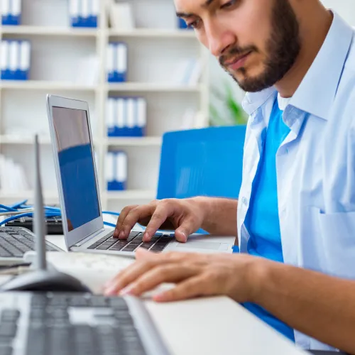 Man working on a laptop at a desk with multiple keyboards and computer equipment in an office setting