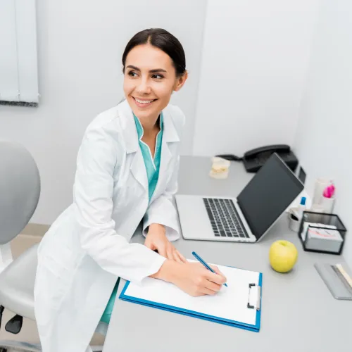 Smiling female dentist in white coat writing on clipboard in dental clinic with equipment and laptop