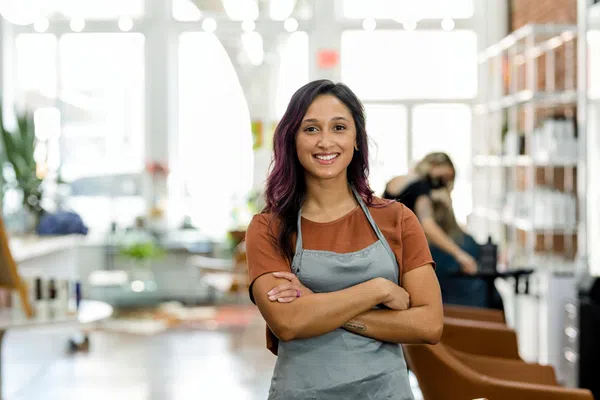 Smiling young woman in gray apron standing with arms crossed inside a bright modern salon.