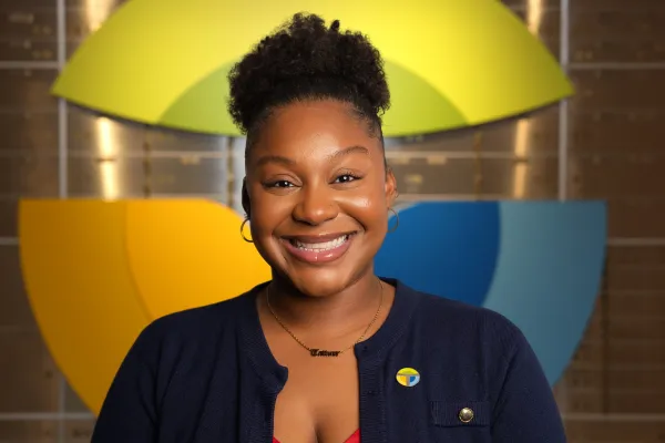 Smiling woman with natural hair wearing a navy cardigan and colorful pin against a geometric yellow and blue background.