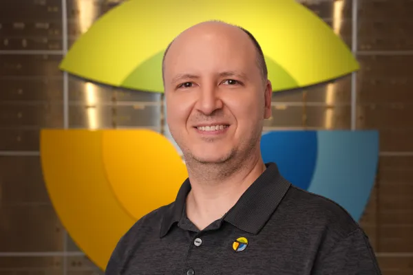 Smiling bald man wearing a black polo shirt with a colorful logo standing in front of a large abstract yellow and blue background.