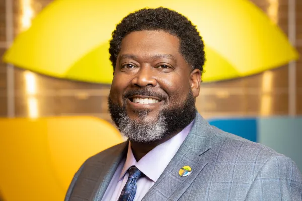 Smiling man with beard wearing gray suit and tie standing against colorful abstract background.