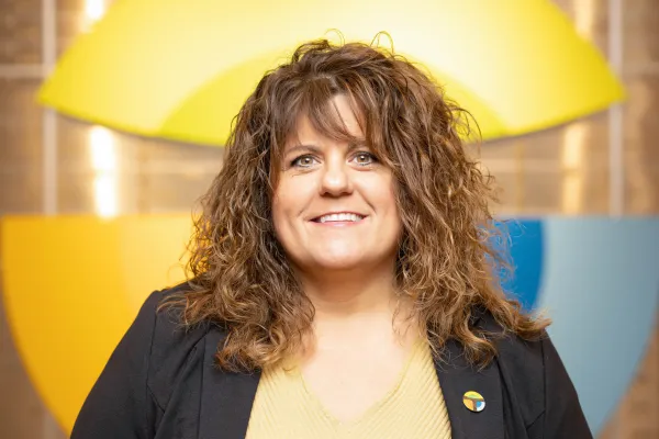 Smiling woman with curly hair, wearing a blazer, standing before a colorful backdrop with yellow and blue shapes.