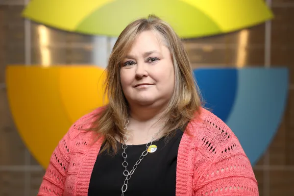 Middle-aged woman with blonde hair wearing a coral cardigan and black top, standing in front of colorful geometric shapes.