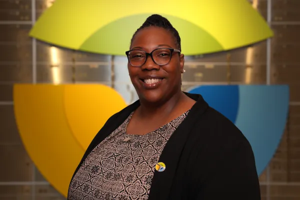 Smiling woman wearing glasses and patterned top stands in front of colorful abstract wall design.