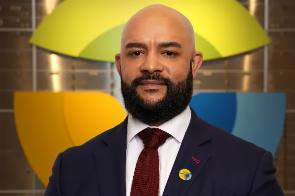 Professional man with beard and bald head in suit and tie standing in front of colorful company logo on wall.