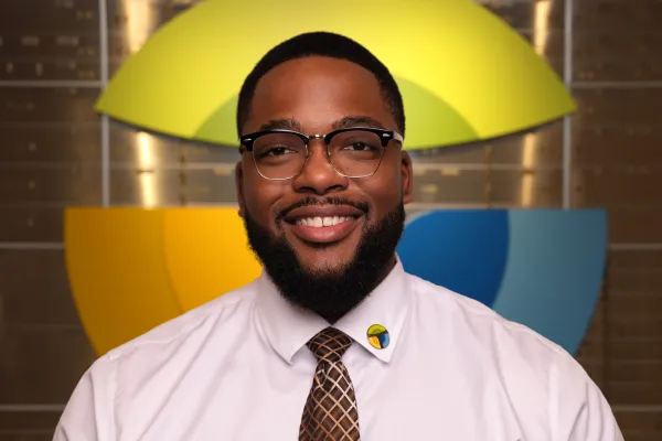 Smiling man in glasses and tie wearing a white shirt with a colorful logo pin and a blurred logo in the background